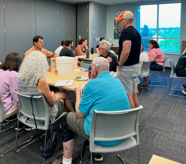 A group of adults sit and stand around tables in a meeting room, engaged in conversation. Some eat food, while others take notes. There is a large window in the background and a plastic container on one table.